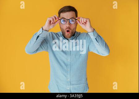 Shocked, touching glasses. Attractive man in blue shirt is against yellow background. Stock Photo
