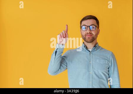 Got an idea, pointing finger up. Attractive man in blue shirt is against yellow background. Stock Photo