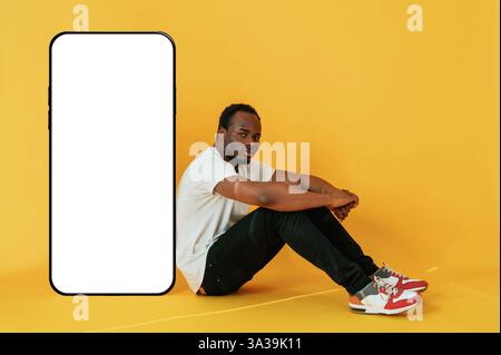 Sitting near to the large smartphone with copy space. Black man is in the studio against yellow background. Stock Photo