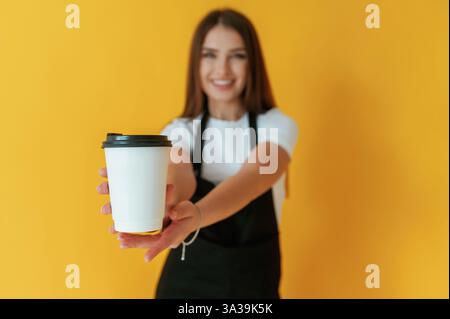 Black and white coffee worker uniform, holding cup of drink. Young woman is against yellow background. Stock Photo