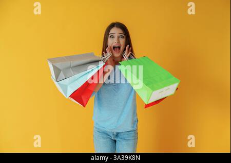 Surprised and anticipated, with shopping bags. Young woman is against yellow background. Stock Photo