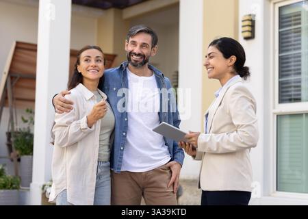 Positive couple of homeowners and young Indian realtor standing outside Stock Photo