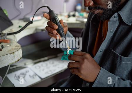 African American man carefully using soldering iron for fixings electronic components in workshop. Tools and components scattered on desk, focused on precise work Stock Photo