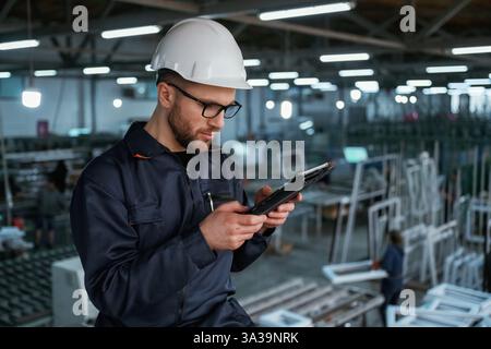Reading the data from tablet. Factory worker is indoors with hard hat. Stock Photo