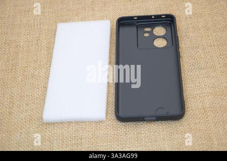 A black plastic phone case lies on its side, next to it is an empty white square piece of foam for packaging the phone case. Stock Photo