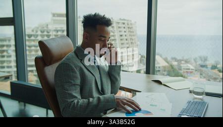 A professional businessman analyzing charts and data while on a phone call in a modern office with large windows and an urban cityscape view. Stock Photo