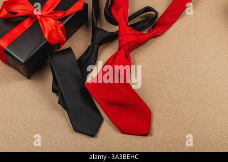 Three stylish ties, black and red, rest next to a beautifully wrapped gift box adorned with a vibrant ribbon. Perfect for father's day or a birthday c Stock Photo