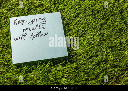 A light blue sticky note with handwritten motivational message is placed on vibrant green grass, reminding viewers to keep going as results will follo Stock Photo