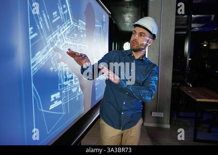 Protective hard hat on head. Man architect in hard hat is near projector with construction plan. Stock Photo