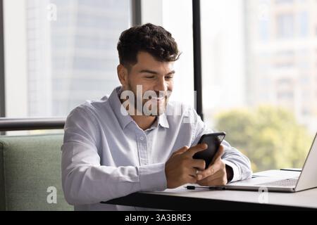 Young man sitting at desk, smiling, looking at smartphone Stock Photo