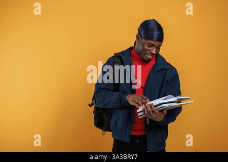 Student with books, backpack and notepad. Handsome black man is in the studio against yellow background. Stock Photo