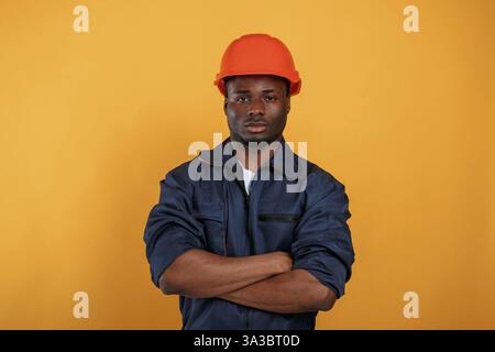 In orange hard hat. Construction worker. Handsome black man is in the studio against yellow background. Stock Photo