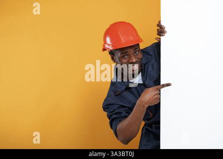 Engineer in hard hat is hiding behind white copy space. Handsome black man is in the studio against yellow background. Stock Photo