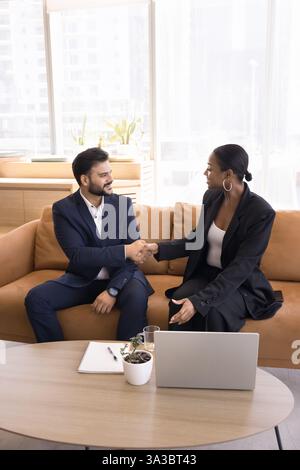 Multiethnic colleagues shake hands seated on couch in office lobby Stock Photo