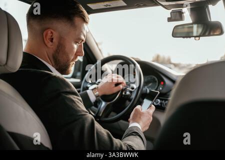 Surfing the internet by using smartphone. Man in black suit is sitting in the car, driving. Stock Photo