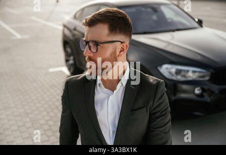 Handsome smart guy in glasses. Businessman in suit is near his black car outdoors. Stock Photo