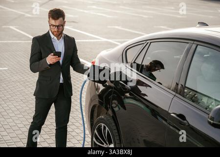 Holding phone in hand while vehicle is charging. Businessman in suit is near his black car outdoors. Stock Photo