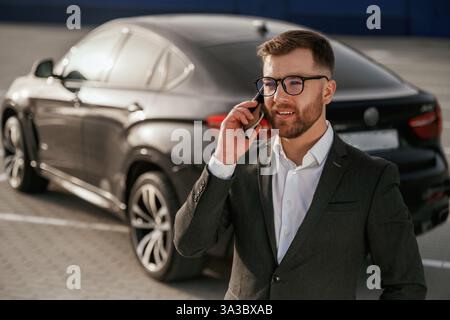 Talking by using phone. Businessman in suit is near his black car outdoors. Stock Photo