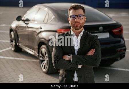 Standing with arms crossed. Businessman in suit is near his black car outdoors. Stock Photo