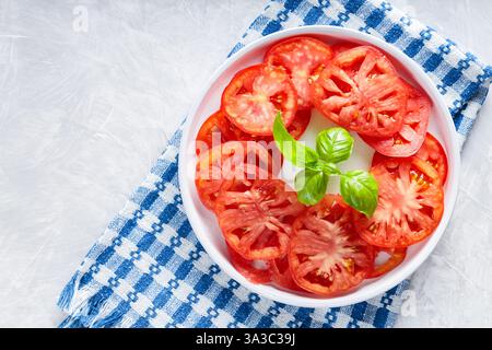 Tomato Slices with Basil Leaves on White Plate, Blue Checkered Napkin, Light Grey Background, Top View Stock Photo