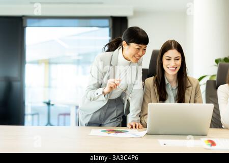 In a contemporary office setting, two women engage in a cheerful conversation, exchanging ideas and smiles while working on a laptop, creating a posit Stock Photo
