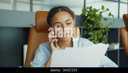 A young professional woman talking on the phone while reviewing documents in a modern office with plants and stylish leather chairs in the background Stock Photo
