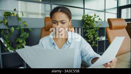 focused young woman analyzing documents at her desk in a modern office, surrounded by charts, a laptop, and office supplies with stylish leather Stock Photo
