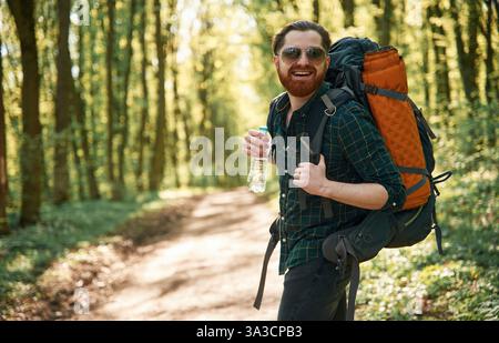 In glasses and with backpack. Bearded man is in the forest at daytime. Stock Photo