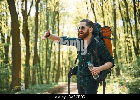 In glasses and with backpack. Bearded man is in the forest at daytime. Stock Photo