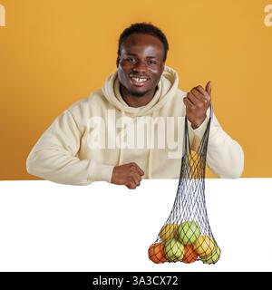 Smiling, holding bunch of fruits against white copy space. Black guy is on yellow background. Stock Photo
