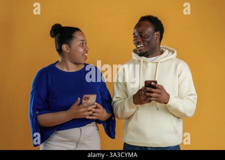 Using smartphones. Happy black couple are together against yellow background. Stock Photo