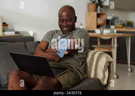 Elderly African American man smiling while checking his smartphone, sitting comfortably on couch with laptop nearby, living room background, insulin monitor on arm Stock Photo