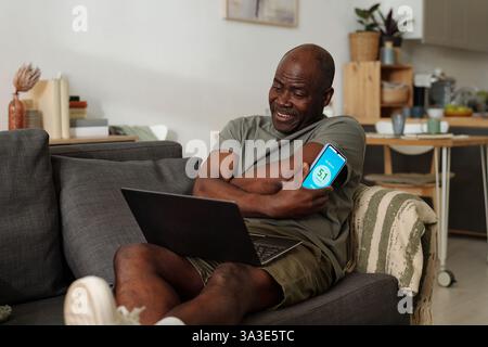 African American man sitting on couch using smartphone and laptop for multitasking, featuring home interior with modern decor in the background Stock Photo