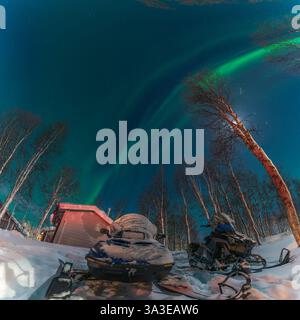 Two snowmobiles resting under a vibrant green aurora borealis illuminate the night sky in Swedish Lappland, surrounded by birch trees Stock Photo