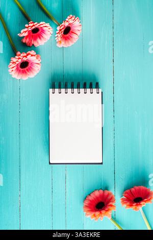 Blank notepad with pink gerbera flowers on a turquoise wooden background. Top view, flat lay, mockup. Stock Photo