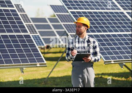 Using walkie talkie and holding notepad. Engineer with photovoltaic solar panels outdoors at daytime. Stock Photo