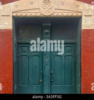 Weathered green double doors with intricate carved details and a decorative stone archway, set against a red brick facade of a historic building. Stock Photo