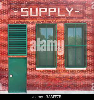 A historic red brick supply building featuring a green metal door, ventilation panel, and security-barred windows, with bold white lettering signage. Stock Photo