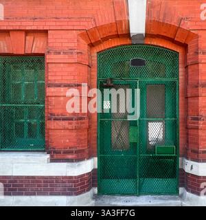 A vintage red brick building with an arched entrance featuring a weathered green metal security gate and window grilles, evoking an industrial era. Stock Photo