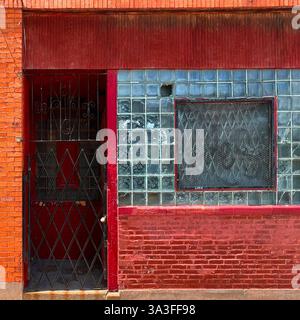 A gated red entrance with a glass block window, covered by a metal security grille, set in a weathered red brick building in an urban environment. Stock Photo