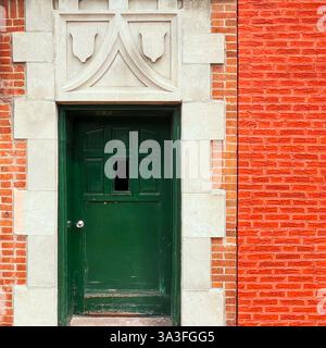Green door with a small window, set within an ornate stone frame featuring decorative shield motifs, surrounded by ornage brick facade Stock Photo
