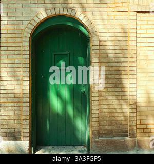 Rustic green wooden arched door set within a yellow brick facade, with dappled sunlight casting shadows in Chicago. Stock Photo
