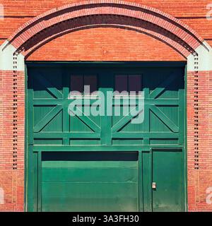 A large green vintage warehouse door with a classic X-brace design, set within a historic red brick building with an arched brickwork. Stock Photo