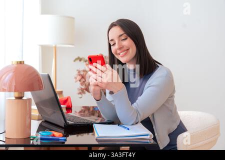 Caucasian smiling student using smartphone while studying at home with laptop on desk Stock Photo