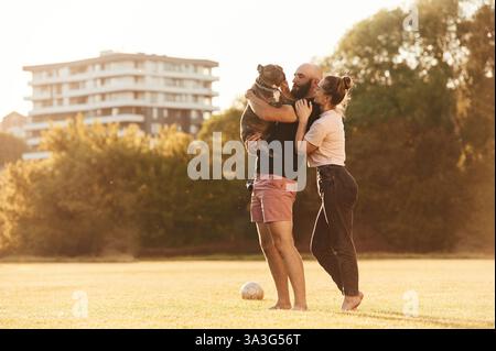 Side view, standing and holding animal. Man and woman are with dog on the field. Stock Photo