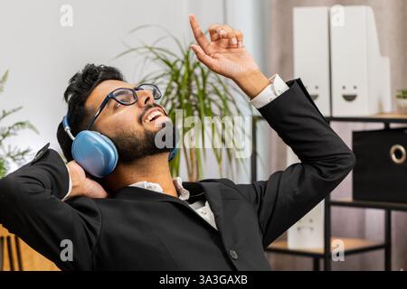 Happy smiling Indian businessman listening music on headphones taking break relaxing dancing leaning on chair after working at office. Male Hindu freelancer in formal suit puts hands behind head Stock Photo