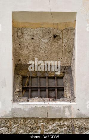 A barred window set in a weathered wall. The wall shows layers of plaster and stone, framed by a newer, smooth white border. The bars are rusty, hinti Stock Photo