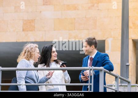 Three business professionals engage in a conversation outdoors, smiling and exchanging ideas while holding smartphones and documents. Modern urban env Stock Photo