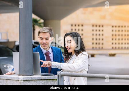 Two business professionals in formal attire discussing work outdoors using a laptop. the woman points at the screen while the man listens attentively, Stock Photo