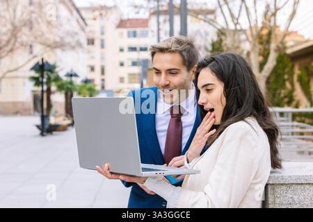 Two young business professionals in formal attire reacting with surprise while analyzing data on a laptop in an outdoor setting. concept of innovation Stock Photo